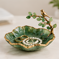 Green leaf-shaped jewelry dish with gold-tone rim and bird branch, displaying rings and a white beaded bracelet