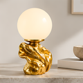 Glowing round globe table lamp on a sculptural gold-tone base displayed on a light tabletop